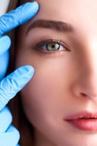 Upper eyelid reduction, double eye lid removal plastic surgery concept. Beautician doctor hands in gloves touching female face skin. Ophthalmologist or oculist checks healthy vision in green eyesight